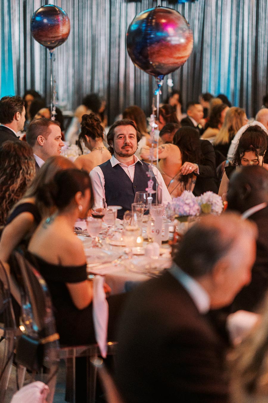 A lively formal event with guests seated around elegantly set tables, featuring dim lighting and colorful planet-themed balloons as decorations.