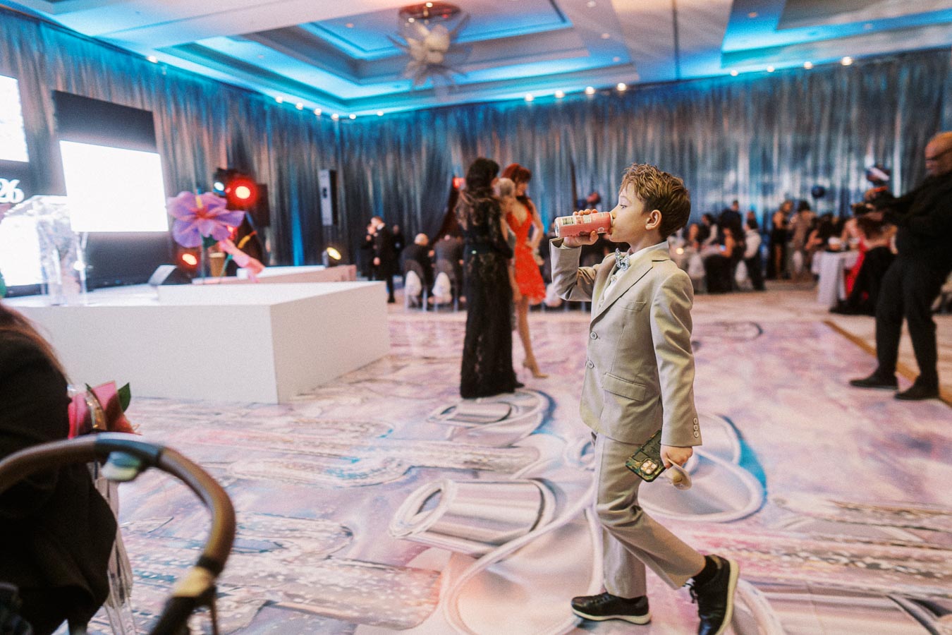 Young boy in a gray suit enjoying a beverage at a stylish event in an elegantly decorated venue with dramatic lighting.