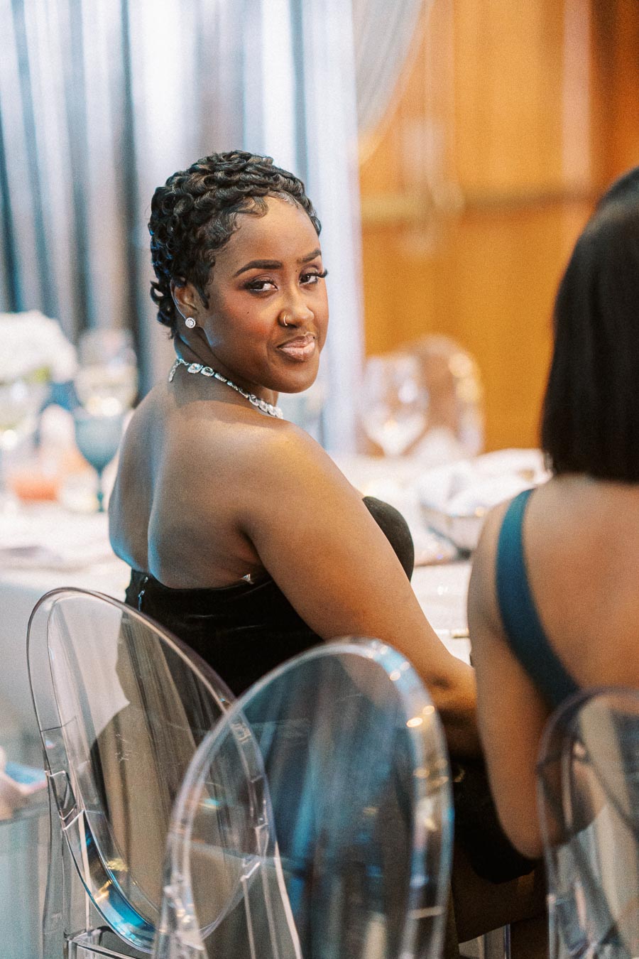 Elegant person at a formal event sitting on a transparent chair, wearing a stylish black dress and a sparkling necklace, looking over shoulder with a smile in a beautifully decorated setting.