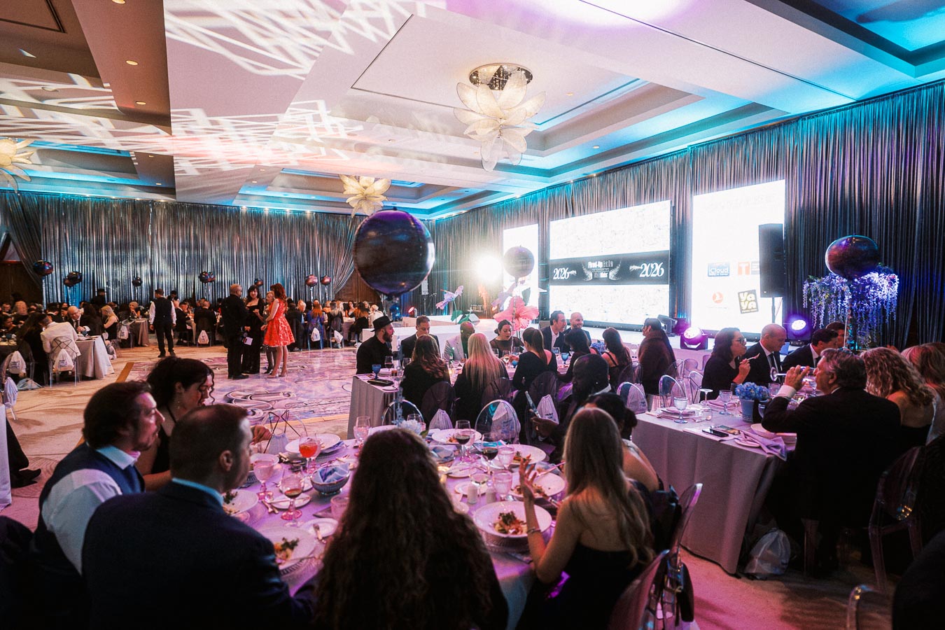 A vibrant and elegant banquet hall filled with guests sitting at round tables during a gala event, featuring decorative lighting, elaborate table settings, and a large screen displaying event branding.