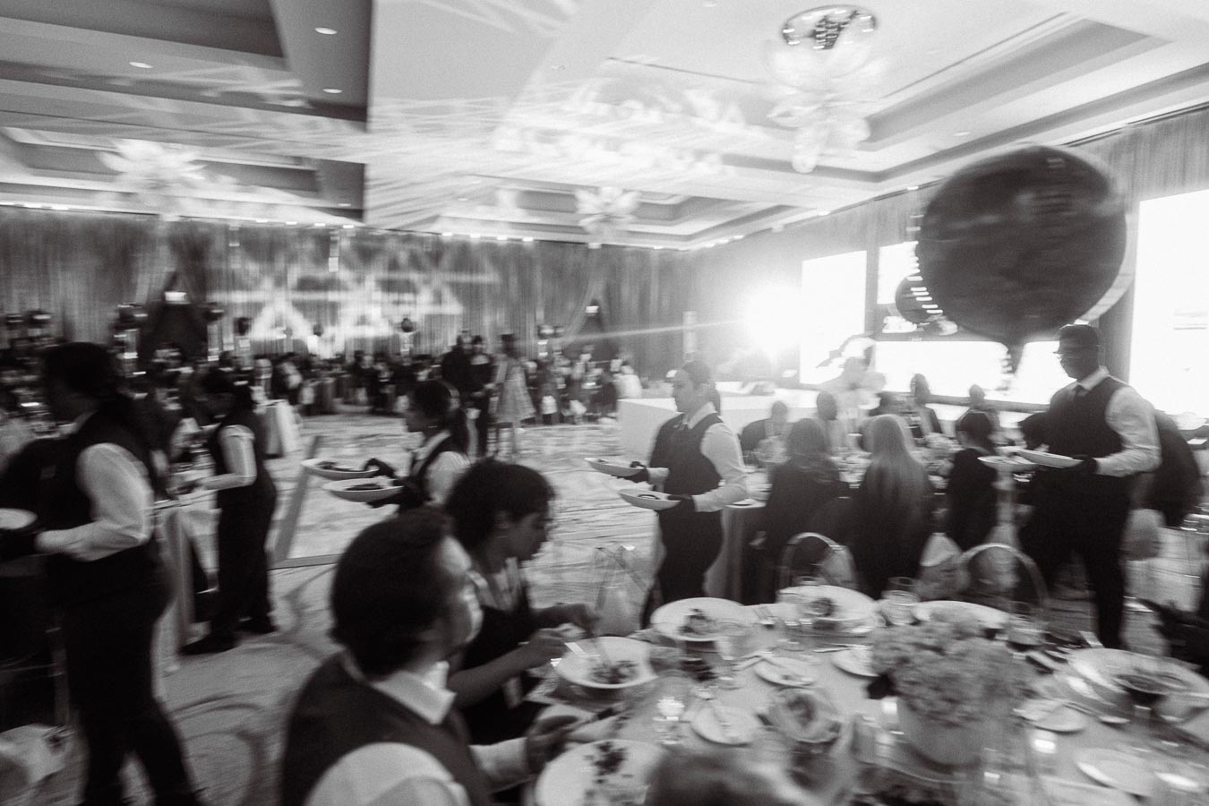 A bustling banquet hall filled with elegantly dressed guests seated at round tables, while waitstaff in uniforms serve food during a formal event, with large decorative elements and ambient lighting creating a festive atmosphere.