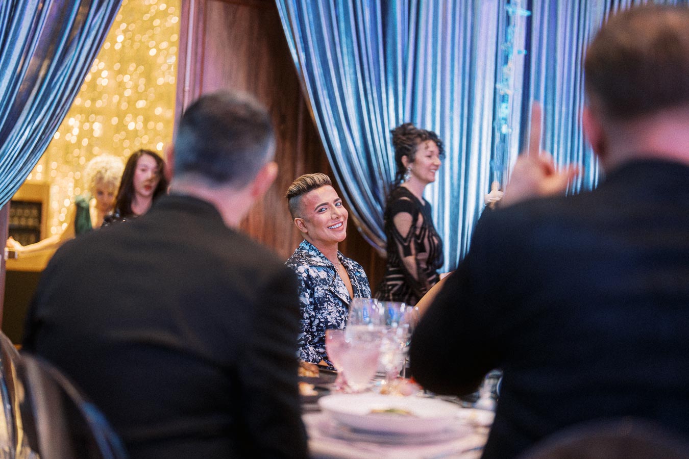 Group of people smiling and conversing at an elegant event with shimmering drapes and elegant attire, focusing on a woman with short hair enjoying a conversation.