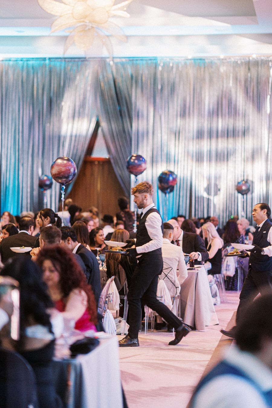 Elegant wedding reception with guests seated at tables, decorated with shimmering silver drapes and floating balloons. Professional waitstaff in formal attire serve food, enhancing the sophisticated atmosphere of the event.