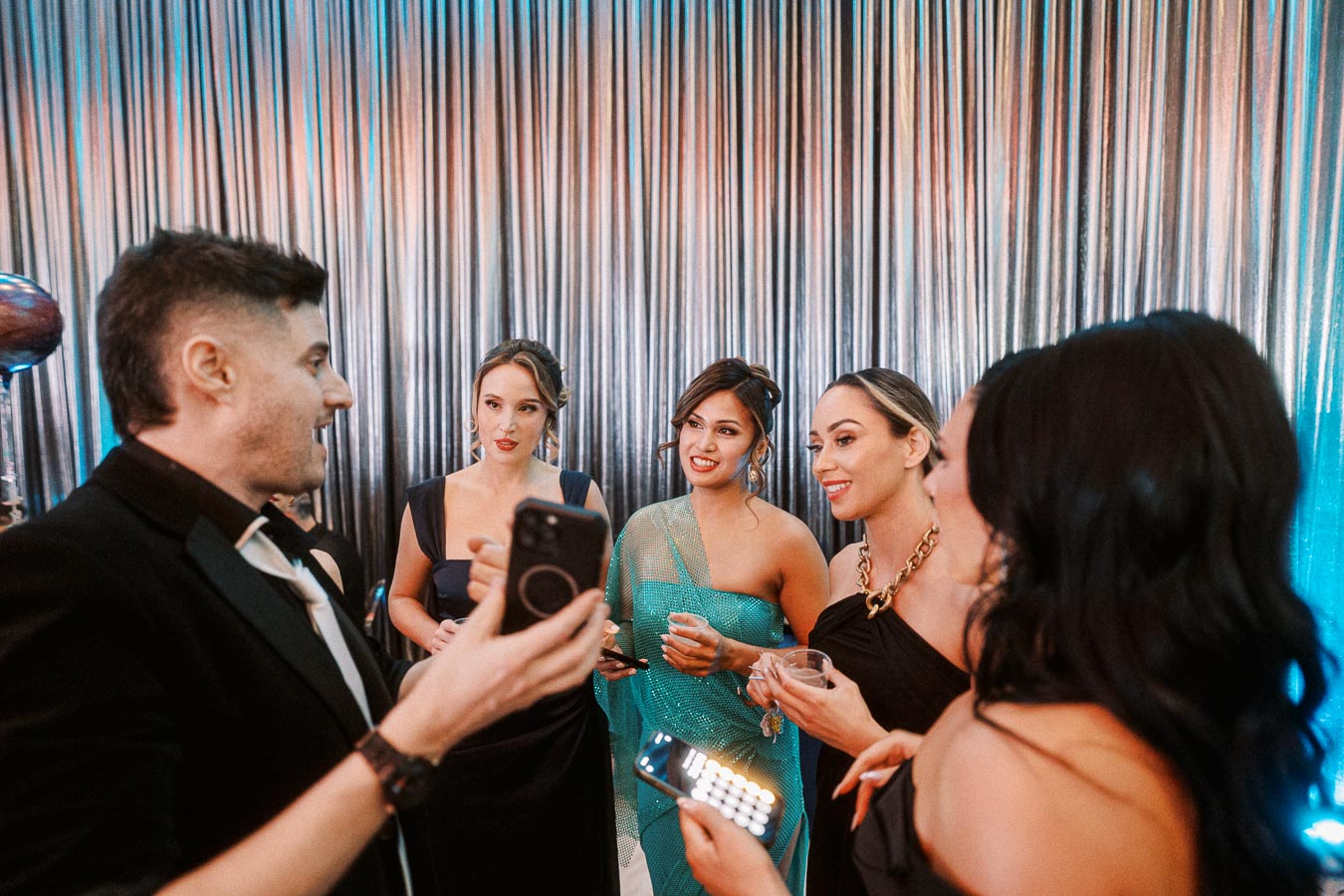 A group of elegantly dressed individuals enjoying a lively conversation at a formal event, with silver drapery in the background, capturing a moment of social engagement and celebration.