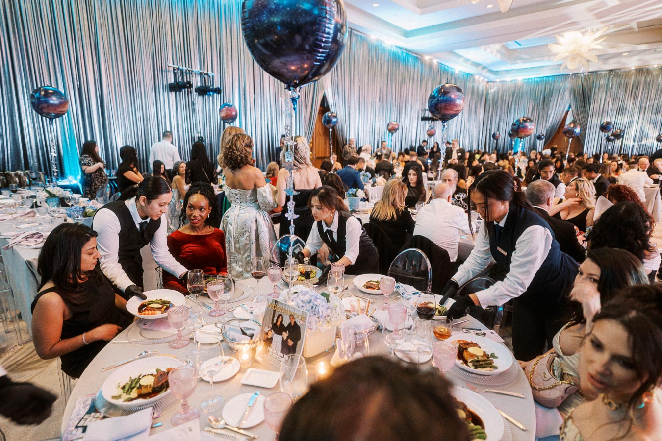 A vibrant banquet hall filled with elegantly dressed guests seated at round tables adorned with fine dining tableware. Waitstaff in formal attire serve gourmet dishes to attendees, while decorative blue and purple balloons add a festive atmosphere. The room is draped in shimmering silver curtains, creating a glamorous setting for the event.