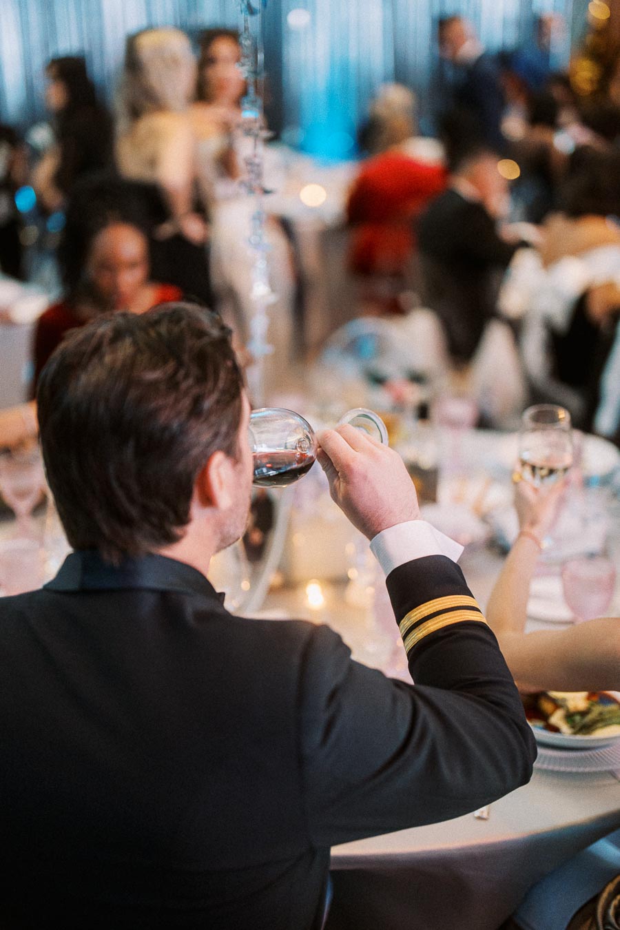 Man in formal attire enjoying wine at an elegant event, surrounded by guests in a beautifully decorated venue.