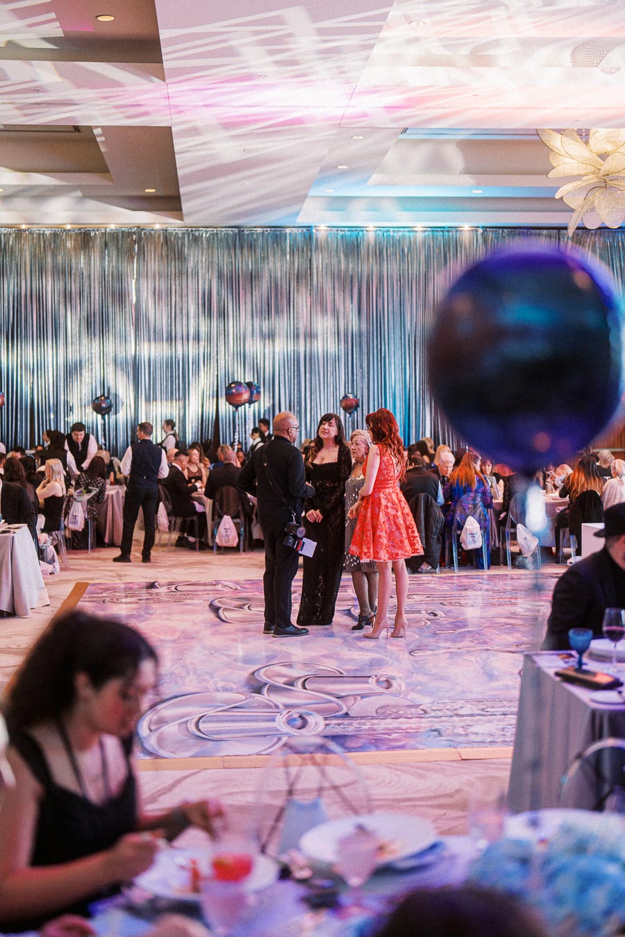 Elegant gala event with guests socializing in an ornately decorated ballroom, featuring shimmering curtains and a patterned floor. Attendees are dressed in formal attire, creating a sophisticated atmosphere.