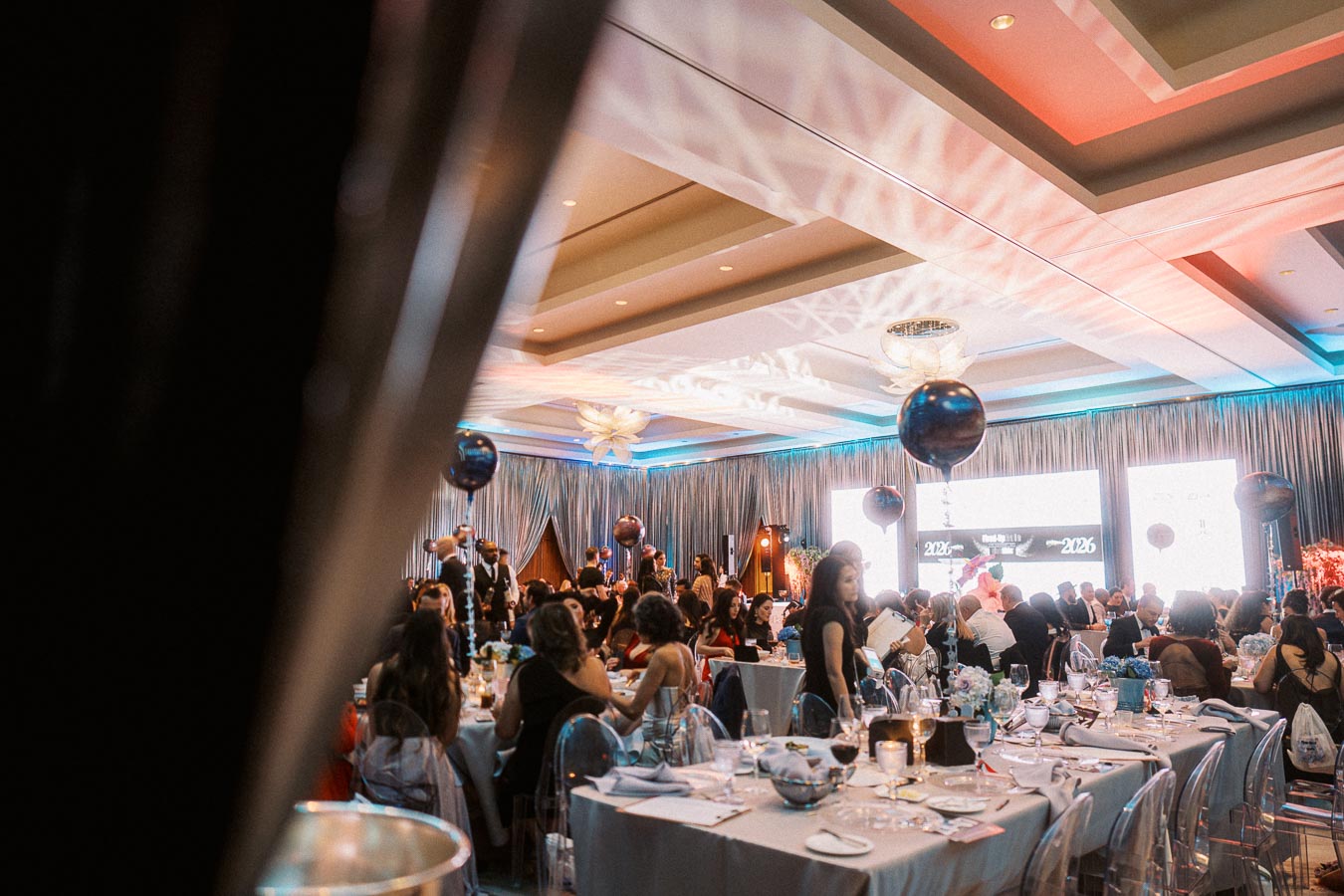 Elegant indoor event with guests seated at decorated tables, featuring dim lighting and blue and silver balloon decorations.