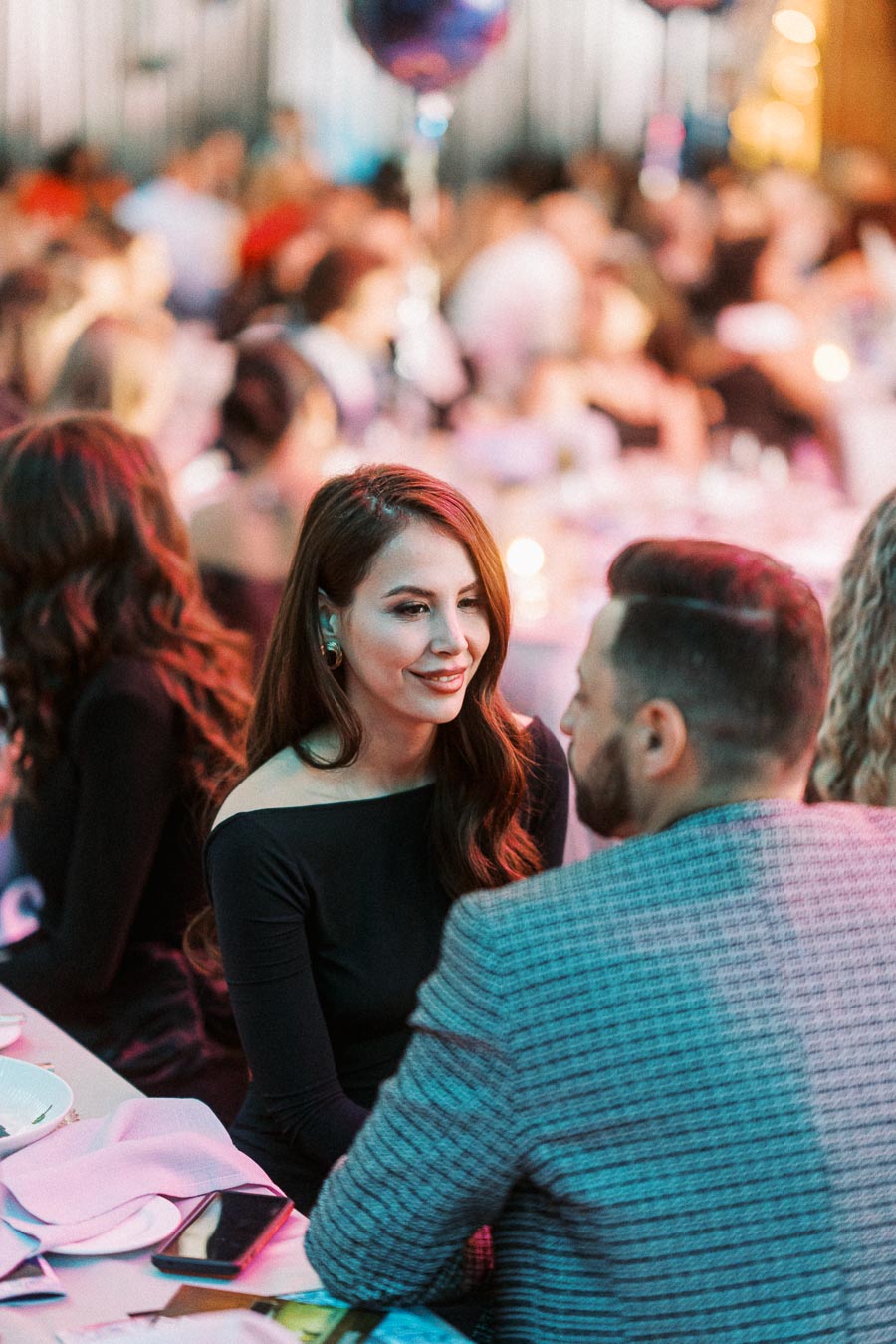 A woman in a black dress engages in conversation with a man in a suit at a lively social event, with a crowd and a decorated venue in the background.