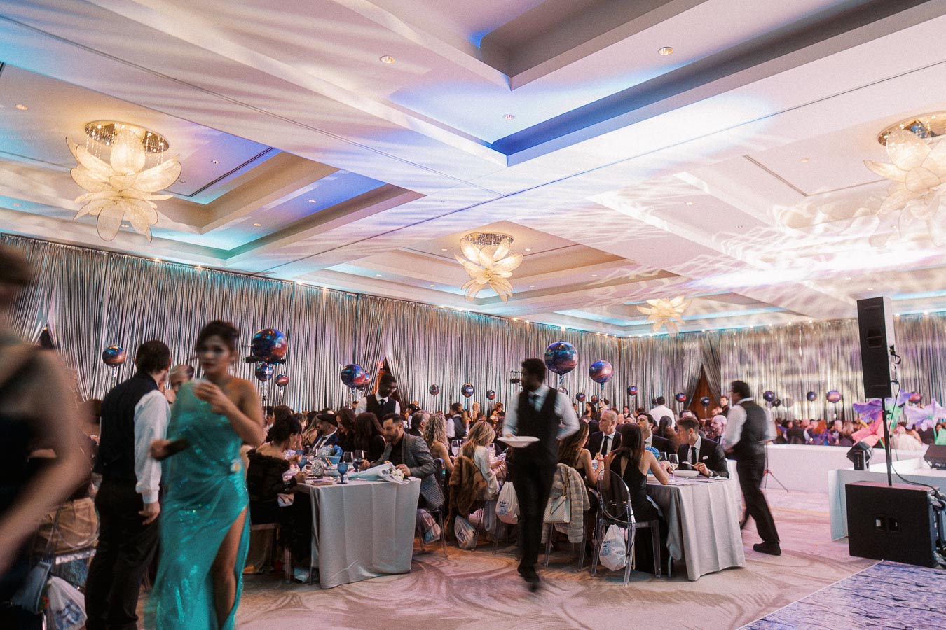 Elegant gala event with guests seated at decorated tables, featuring a shimmering ceiling and ambient lighting, creating a luxurious atmosphere. Waitstaff serve attendees, enhancing the event's formal and festive setting.