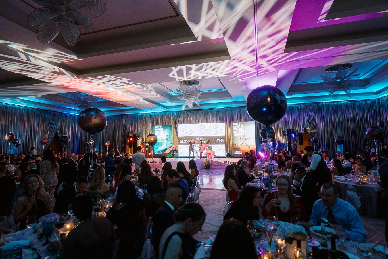 Luxurious gala event with elegantly dressed attendees seated at tables, ambient lighting, decorative balloons, and a colorful stage featuring vibrant lighting effects.