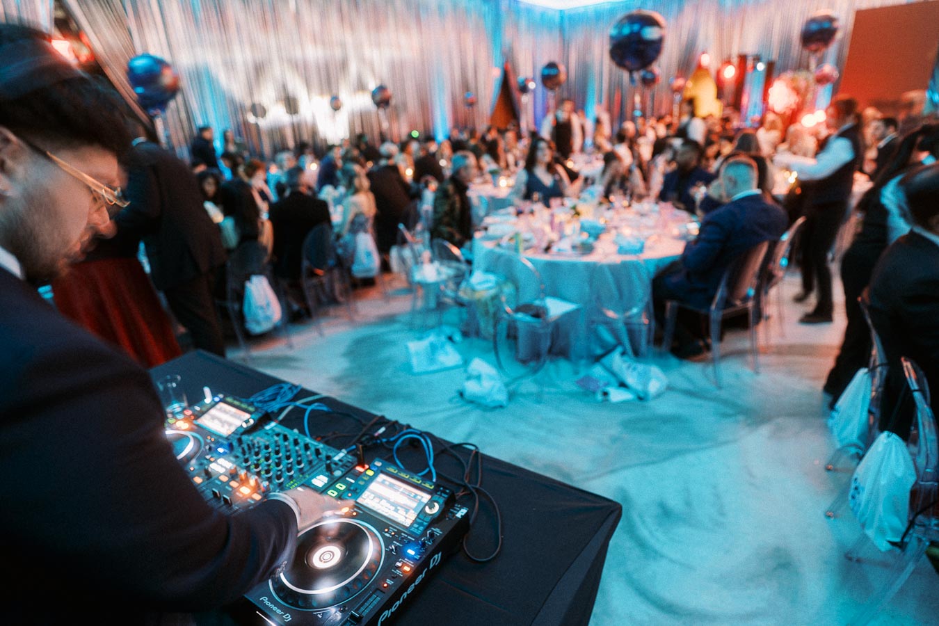 DJ mixing music at a lively party with guests seated at round tables, colorful lighting, and decorative balloons.