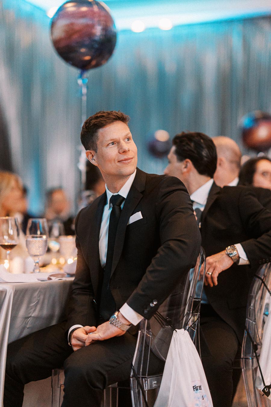A man in a black suit sits attentively at a formal event, surrounded by elegantly set tables and a softly lit atmosphere. He appears engaged, with a balloon floating nearby, adding a festive touch to the decor.
