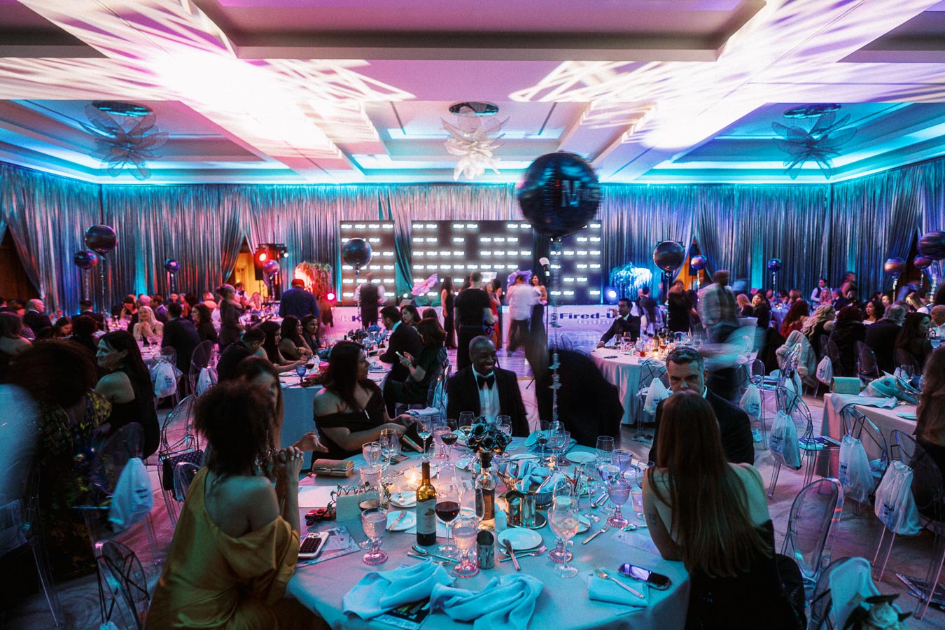 Elegant gala event in a lavishly decorated ballroom with guests seated at round tables enjoying a formal dinner. The ambiance is enhanced with colorful lighting, balloons, and a lit stage in the background.