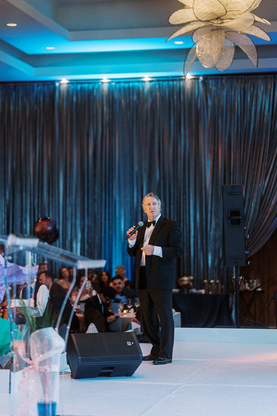 A man in a suit and bow tie holding a microphone on stage at a formal event, with blue lighting and an audience in the background.