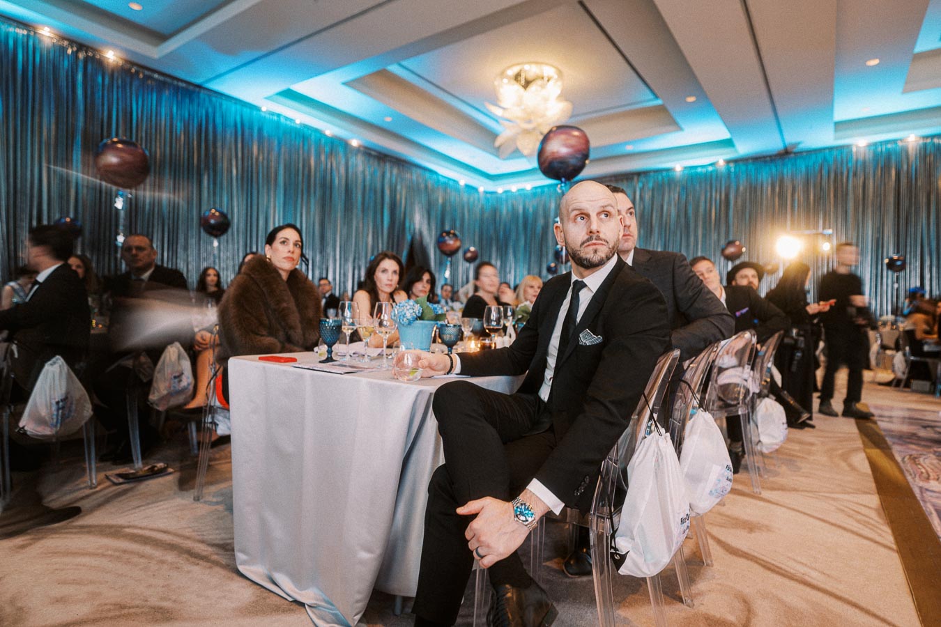 A formal evening event with attendees seated at elegantly decorated tables, featuring blue lighting, balloons, and a sophisticated atmosphere.