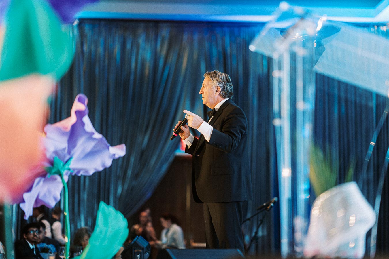 Man in a suit speaking into a microphone at a formal event, with large colorful flowers in the foreground and blurred audience in the background.