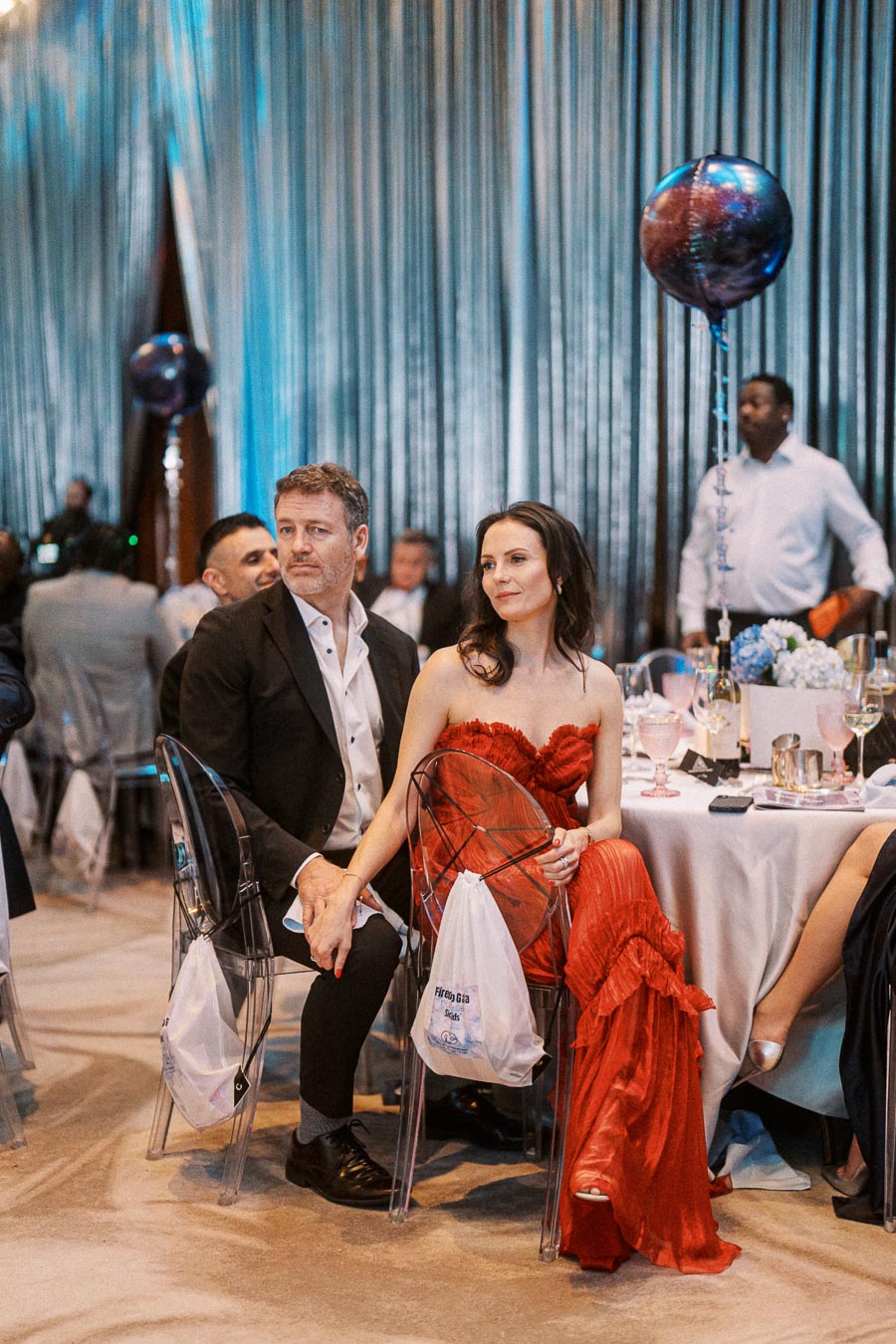 Elegant couple holding hands at a formal event with a red dress and black suit, surrounded by a beautifully decorated table setting, silver curtains, and colorful balloons in the background; sophisticated gala atmosphere.