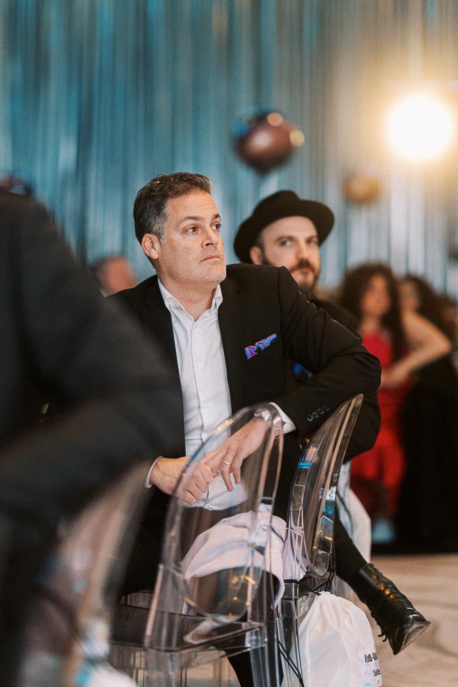 Man in a suit attentively listening at a formal event, seated on a transparent chair, with people in the background and decorative lighting.