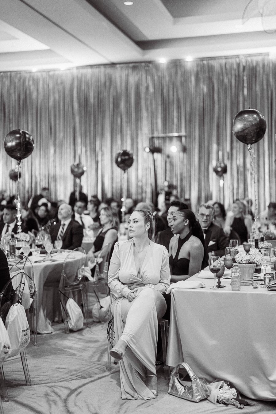 A black and white photograph of a formal event, showcasing elegantly dressed guests seated at round tables adorned with floral centerpieces and wine glasses. The room is decorated with metallic streamers and balloons, creating a festive atmosphere.