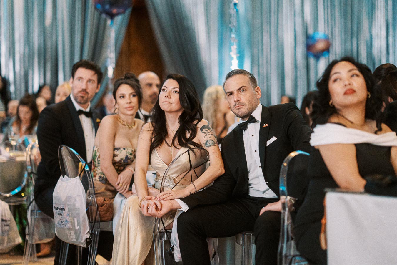 A group of elegantly dressed individuals sitting attentively at a formal event, surrounded by shimmering decor and draped curtains.