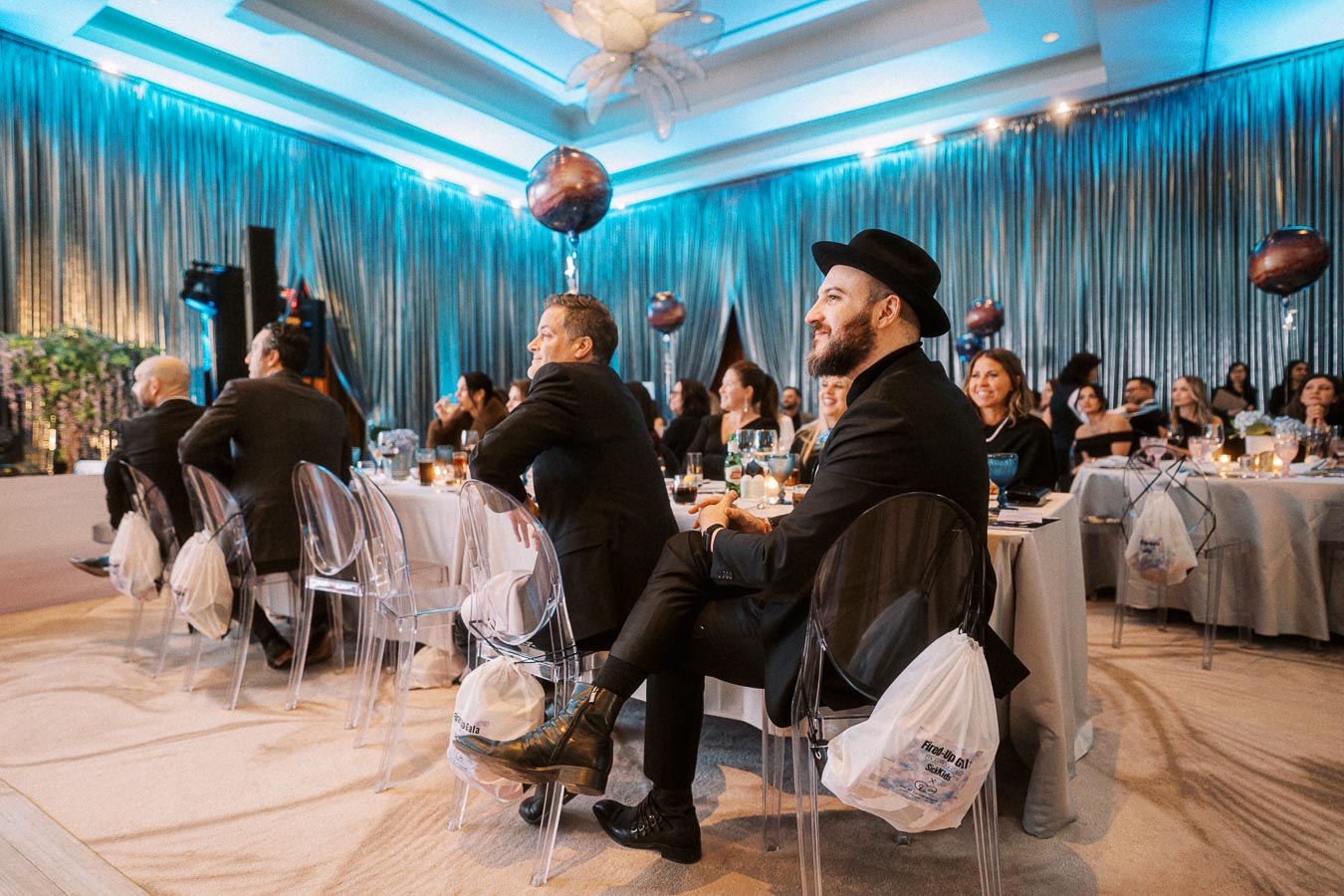 A group of people attentively seated at a formal event in a beautifully decorated venue with elegant drapes and ambient lighting.