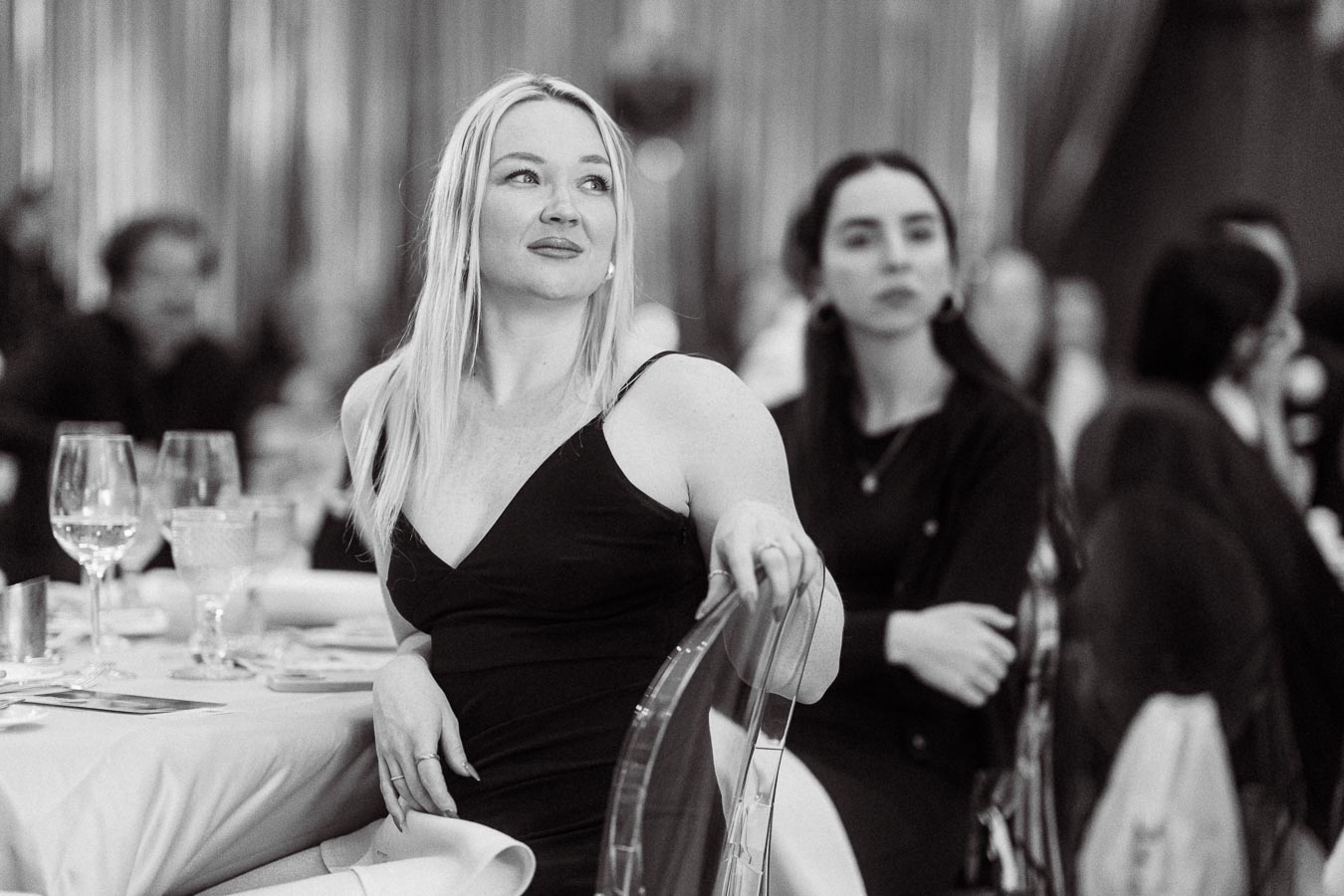 Black and white photo of a woman sitting at a formal event, holding a chair with a thoughtful expression, with another person in the background.