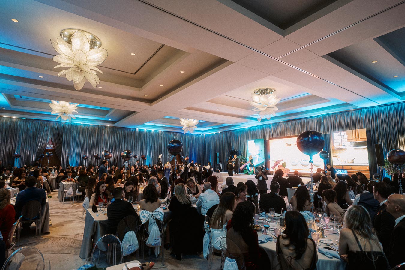 Elegant gala event with attendees seated at round tables in a beautifully decorated ballroom with blue ambient lighting and hanging chandeliers. A stage at the front displays a presentation, with people enjoying the social gathering and live entertainment.