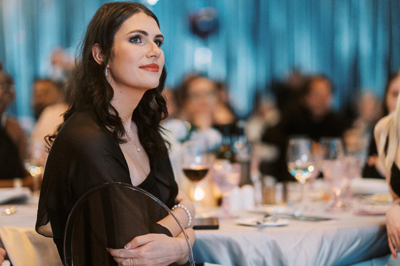 A woman in formal attire attentively listens at a beautifully set table during a sophisticated event, with a blurred background of attendees and elegant decor.