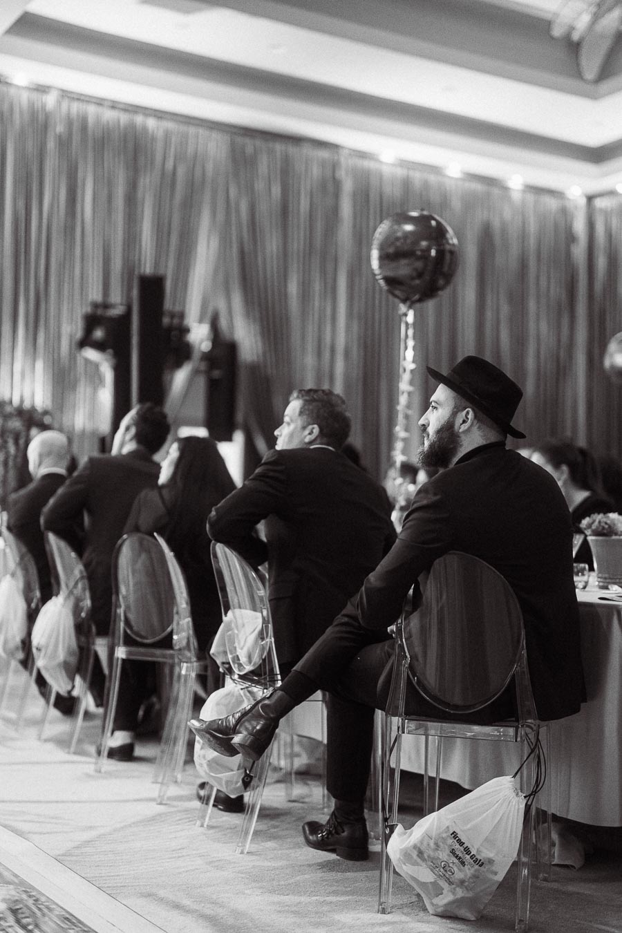 Black and white image of elegantly dressed attendees seated at a formal event, with transparent chairs and decorative balloons in a sophisticated venue setting.