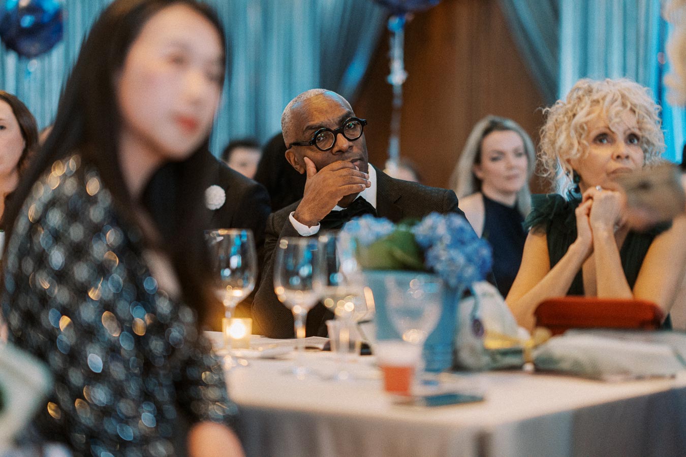 A diverse group of elegantly dressed people attentively listening at a formal event, with soft lighting and blue floral decorations in the background.