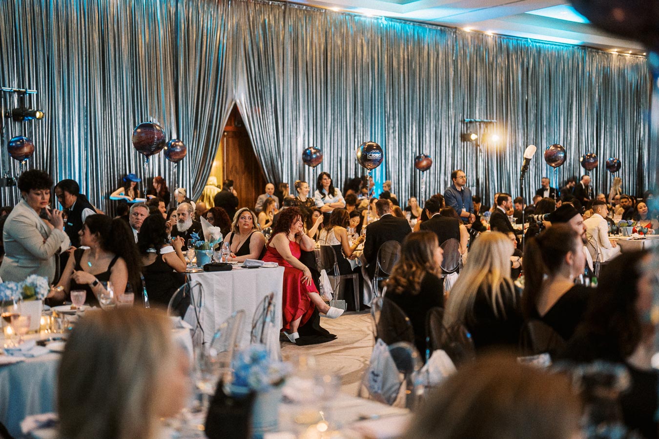 A lively crowd attending a formal event with elegant silver drapery and decorative balloons, featuring seated guests at round tables set with floral centerpieces and dining settings in a sophisticated banquet hall.