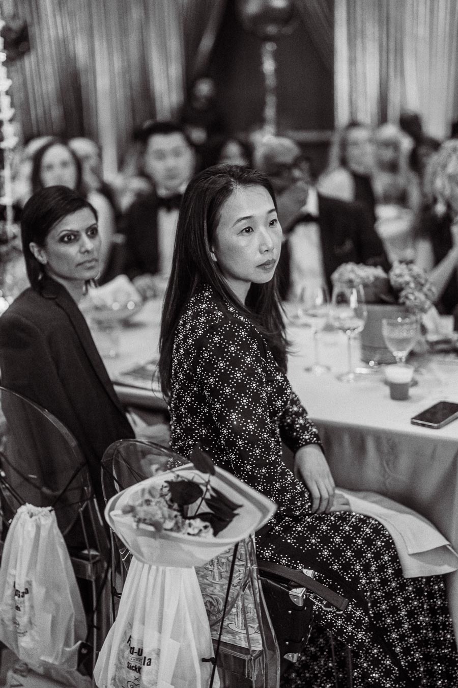 Black and white photo of a formal dinner event with attendees seated at a table, focusing on a woman in a patterned dress and another in a suit, with elegant decor and a bouquet on a chair, capturing a sophisticated gathering atmosphere.