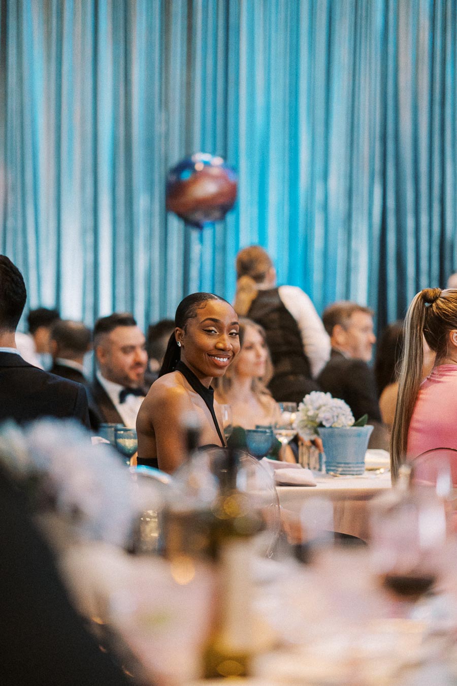 A woman in an elegant black dress smiles at a formal event, surrounded by other guests seated at a beautifully decorated table with blue accents.