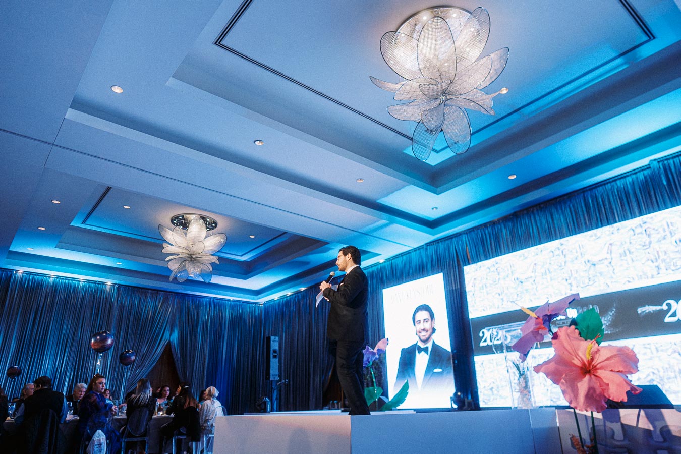 A speaker in a suit addresses an audience at an elegant blue-lit event with decorative flower chandeliers, featuring a large screen showcasing an image of a man in formal attire, suggesting a formal presentation or award ceremony.