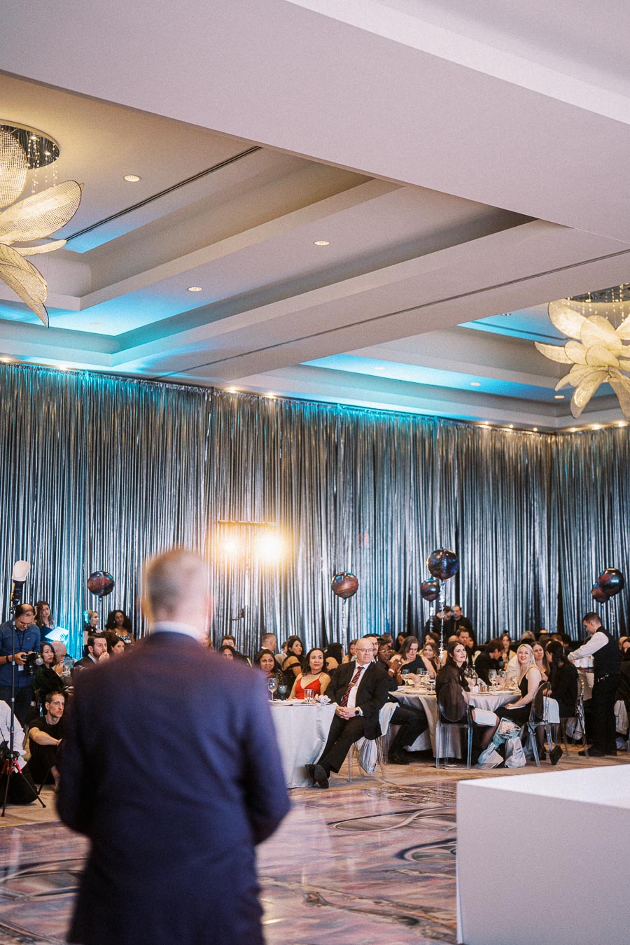 A speaker addresses a seated audience at a formal event with elegant decor and vibrant lighting in a banquet hall.