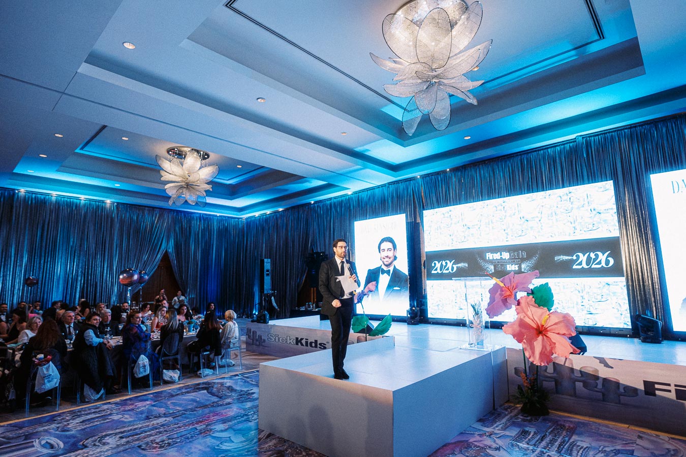A man in a suit stands on stage at a formal event held in a luxurious venue with blue lighting and elegant chandeliers. A large audience watches attentively, and the backdrop displays year 2026 alongside a floral decoration, indicating a gala or fundraiser for SickKids.