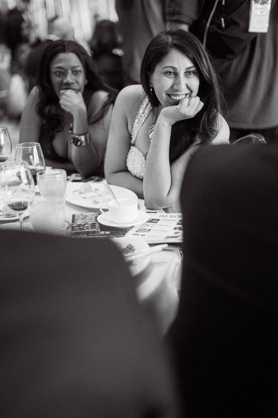 Black and white photo of two women smiling and enjoying a conversation at a formal event, seated at a table with glasses and dinnerware