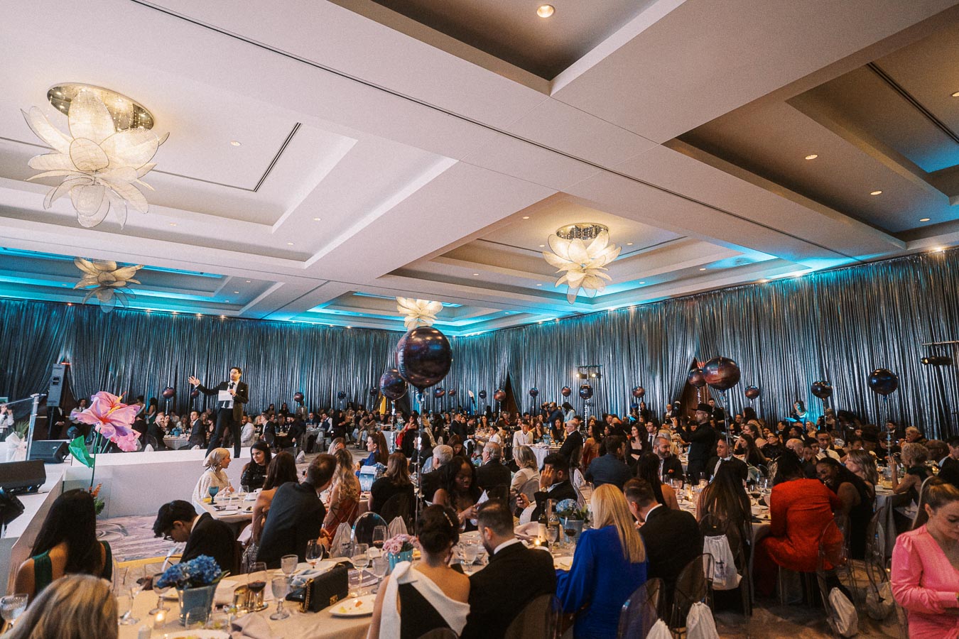 Lavish corporate event in an elegantly decorated ballroom with a presenter on stage, large audience seated at round tables, and stylish lighting creating a sophisticated ambiance.