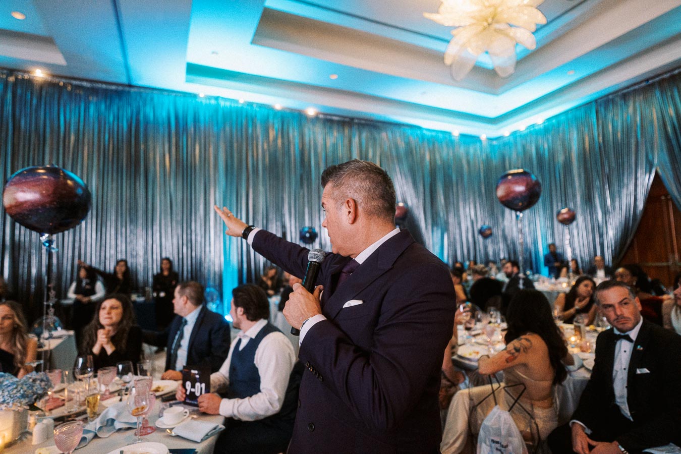 A man in a suit holding a microphone speaks to an elegantly dressed audience at a formal event, surrounded by orb-shaped balloons and soft blue lighting, in a luxurious banquet setting.