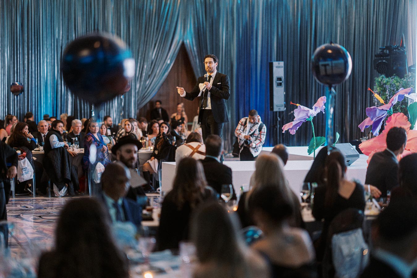 A speaker in a suit delivers a presentation on stage at a formal event, surrounded by an attentive audience seated at tables, with decorative flowers and balloons in the venue.