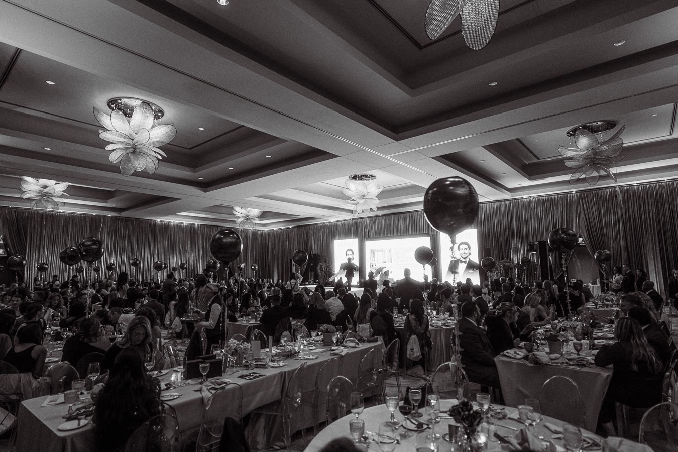 Black and white photo of a formal banquet or gala event in a spacious ballroom, featuring elegantly set tables filled with guests, surrounded by large decorative balloons and dramatic ceiling lighting.