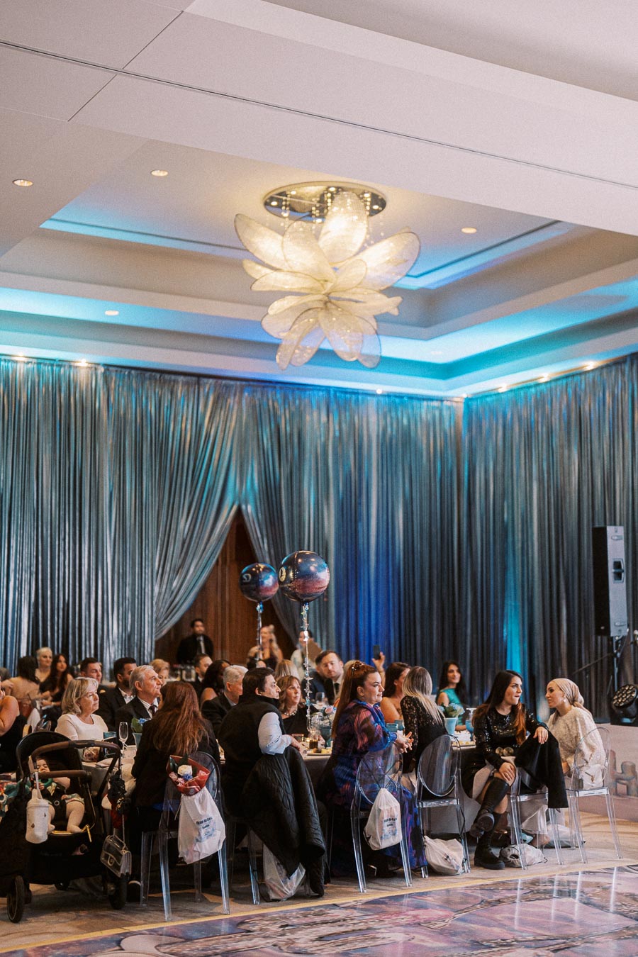 Elegant banquet hall with guests seated at decorated tables, featuring a stunning chandelier and blue-lit drapery, creating a sophisticated atmosphere for a formal event.