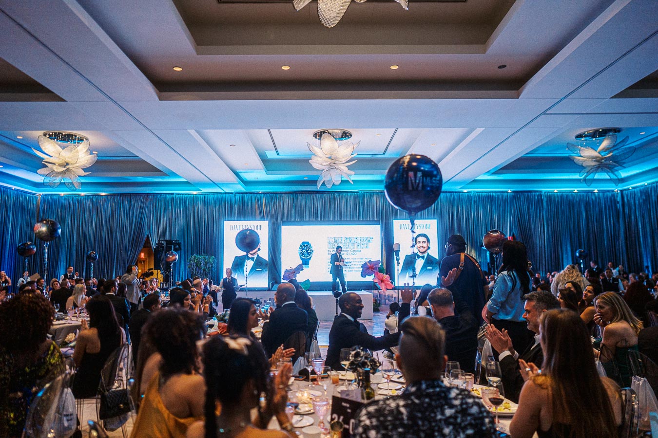 Elegant ballroom event with a large audience attentively watching a speaker on stage with a dynamic presentation. The stage is set with sophisticated lighting, and the atmosphere is lively with attendees seated at round tables adorned with decorations.