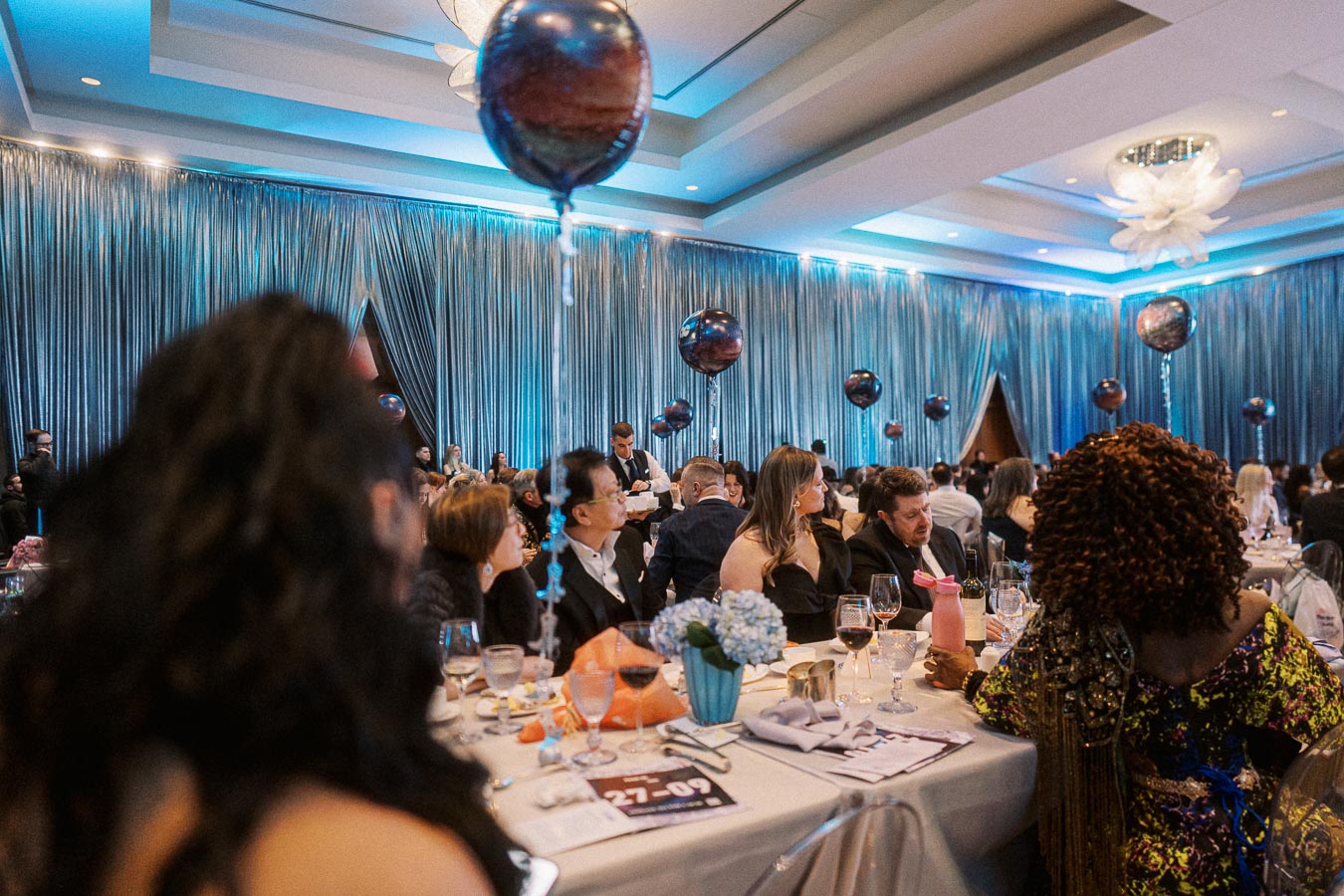 Elegant event with guests seated at decorated tables, featuring metallic balloons and soft blue lighting in a stylish venue.