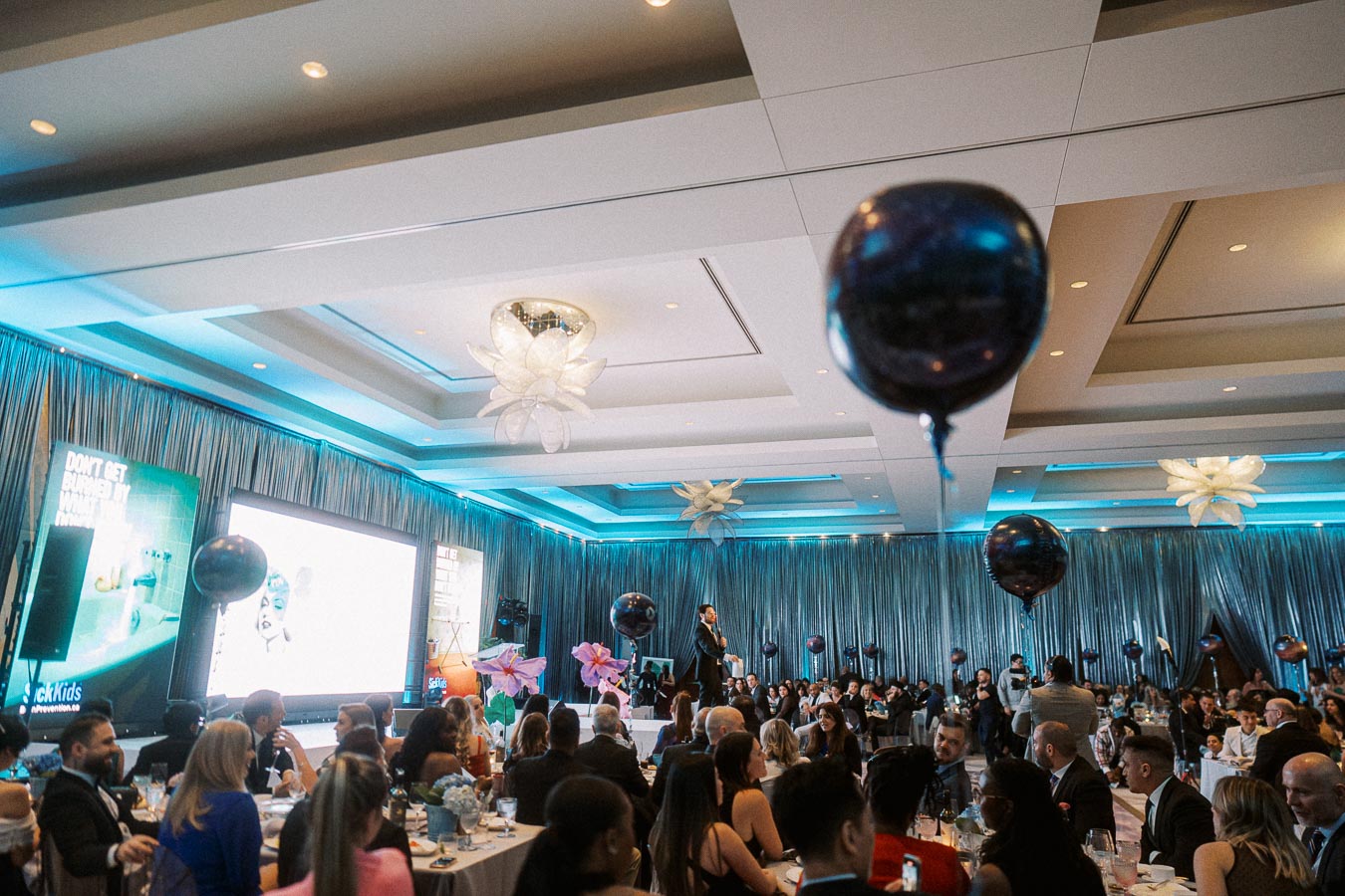 Elegant corporate event with attendees seated at tables, decorated with dark balloons, under stylish lighting. A speaker presents on stage near large screens and vibrant floral arrangements in a sleek, modern venue.