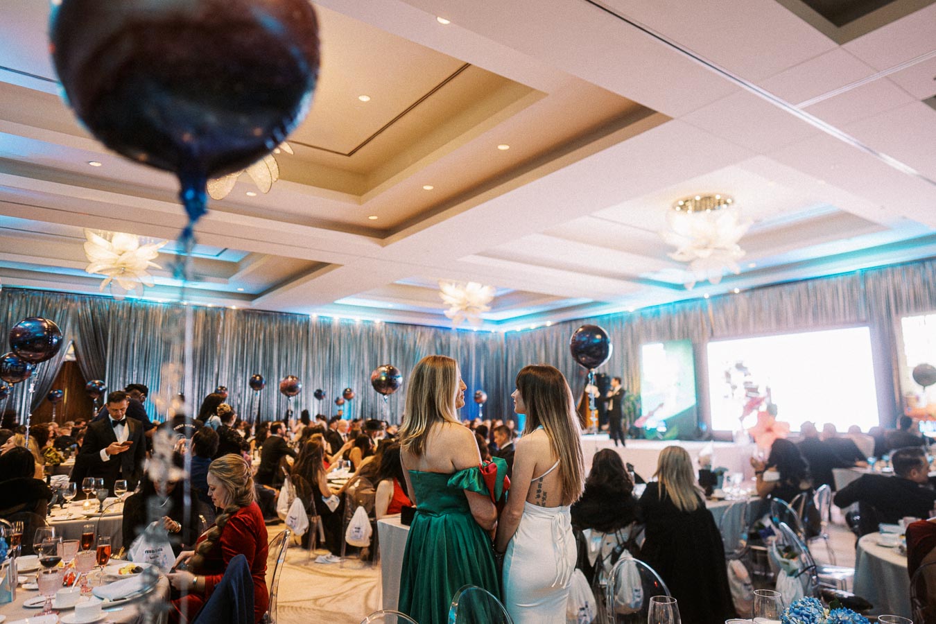 A luxurious banquet hall decorated for an elegant evening event, featuring round tables with guests dressed in formal attire, vibrant balloons, and a stage with speakers and presentations.