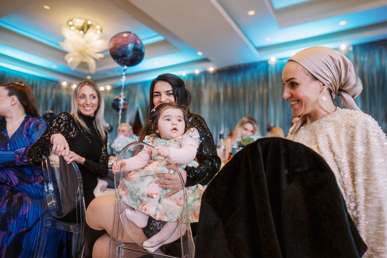 A group of women at a festive gathering, with one holding a baby in a floral dress. The room is decorated with balloons and elegant lighting, creating a joyful and celebratory atmosphere.