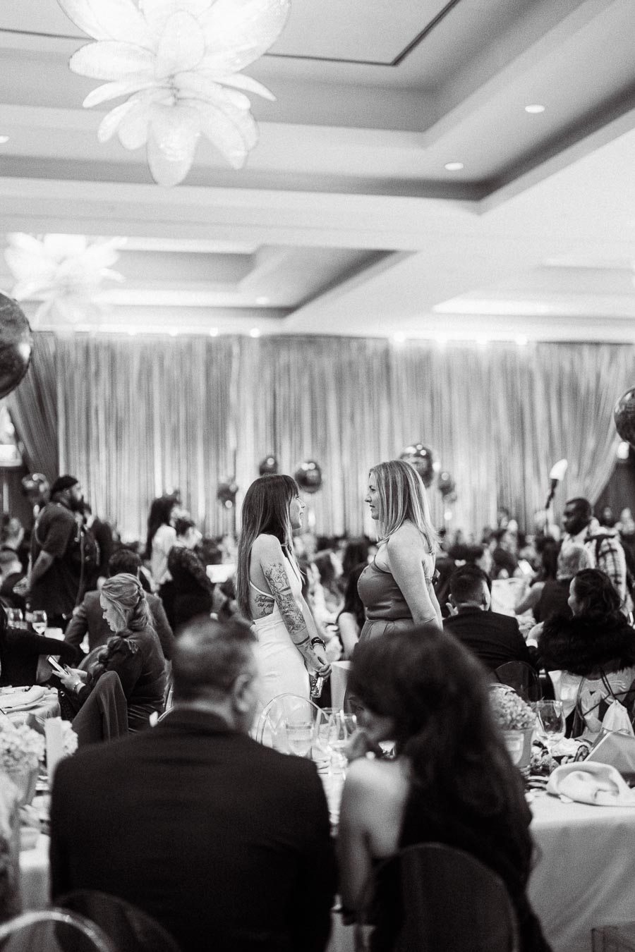 Black and white image of a formal event, featuring elegantly dressed guests seated at tables. Two women are engaged in conversation in the center, surrounded by a festive atmosphere with balloons and lavish decor. A luxurious venue with ambient lighting adds to the sophisticated setting.