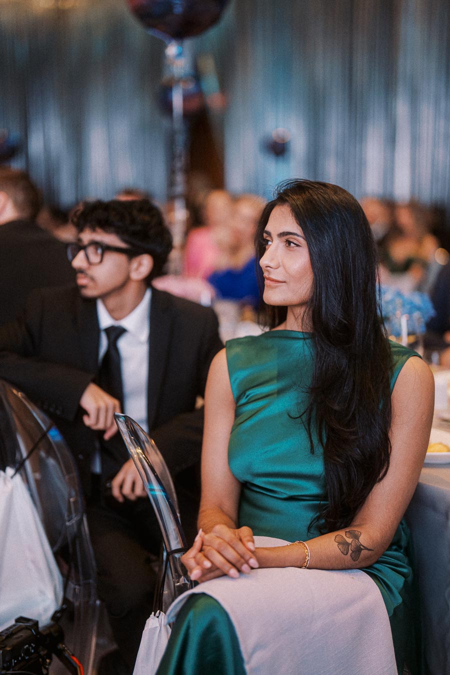 Two elegantly dressed individuals attending a formal event, with a woman in a green dress featuring a floral tattoo on her arm and a man in glasses and a suit, seated at a table in a sophisticated setting.